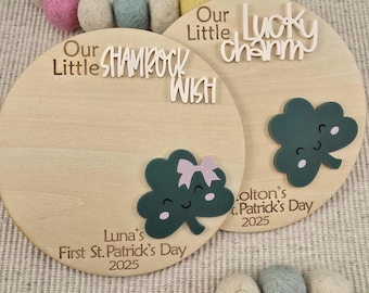 May include: Two round wooden signs with the text "Our Little Shamrock Wish" and "Our Little Lucky Charm" on them. Each sign has a green shamrock with a smiling face and a pink bow. The signs are decorated with small, colorful balls.