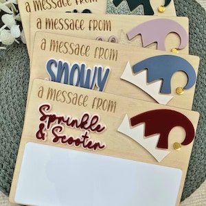 May include: Wooden message boards with elf hat designs in various colors, including green, purple, blue, and red. The boards feature the text "a message from" and names like "Snowy" and "Sprinkle & Scooter."
