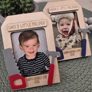May include: Two wooden photo frames with the text "DAD'S LITTLE HELPER" and "PAPA'S LITTLE HELPERS". Each frame holds a child's photo and decorative tools. The text "HAPPY FATHER'S DAY 2025" is also visible.