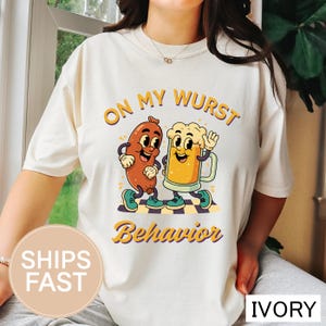 Può includere: T-shirt color avorio con la scritta "ON MY WURST Behavior" e un'illustrazione a cartoni animati di una salsiccia e un boccale di birra. La salsiccia e il boccale di birra camminano insieme su un pavimento a scacchi. La maglietta ha uno scollo tondo.