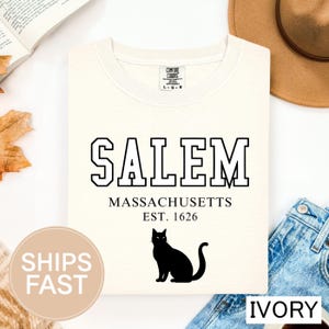 May include: Ivory-colored sweatshirt with the word "SALEM" in bold, black, block letters. Below, the text reads "MASSACHUSETTS EST. 1626." A black cat silhouette is printed below the text. The shirt is a Comfort Colors brand.