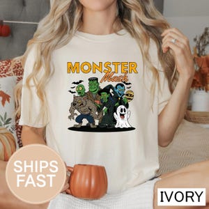 May include: Ivory-colored t-shirt featuring a cartoon graphic of various monsters, including a werewolf, Frankenstein's monster, and a vampire. The text "MONSTER MASH" is printed above the characters in yellow and orange. The shirt is made of a soft material.