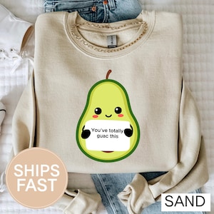 May include: A sand-colored sweatshirt featuring a cartoon avocado character holding a sign that reads "You've totally guac this." The avocado is light green with a brown stem and black eyes. The sweatshirt has a crew neck and long sleeves.