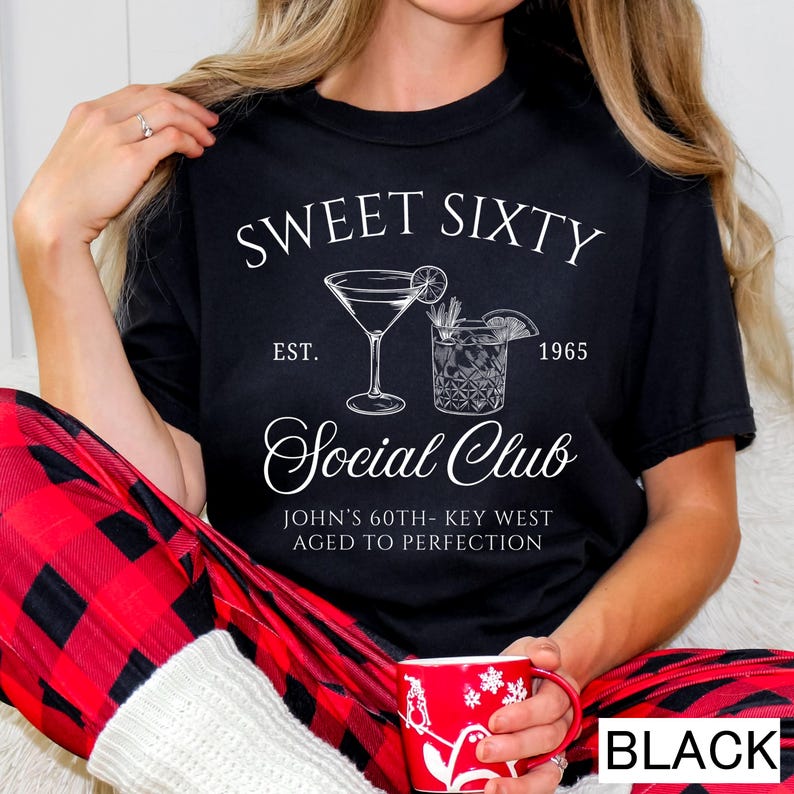 A woman wearing a black t-shirt with a graphic design of a martini glass and a cocktail, along with text that says "Sweet Sixty" and "Est. 1965". She is sitting on a couch with a red mug in her hand.