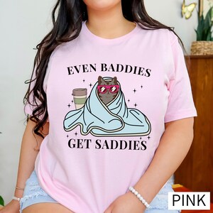 a woman wearing a pink shirt that says even baddies get saddles