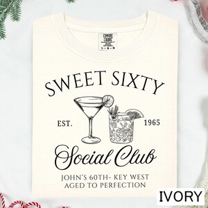 a white t-shirt with a graphic design that includes a cocktail glass and a text that reads "Sweet Sixty Social Club John's 60th - Key West Aged to Perfection".
