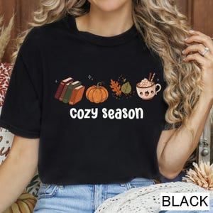 May include: A black short-sleeve t-shirt featuring a graphic print of three stacked books, an orange pumpkin, autumn leaves, and a mug of hot cocoa with whipped cream. The text "cozy season" is printed in white below the graphic elements.