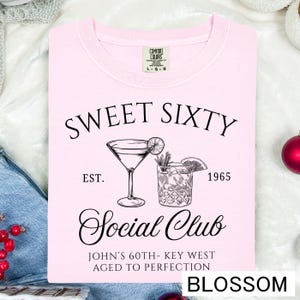 a pink t-shirt with a graphic design and text, including a cocktail glass and the text "Sweet Sixty" and "Est. 1965 Social Club John's 60th - Key West Aged to Perfection".