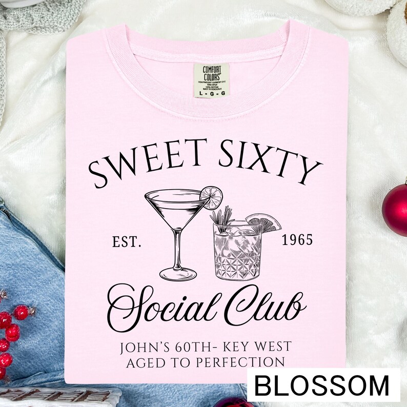 a pink t-shirt with a graphic design and text, including a cocktail glass and the text "Sweet Sixty" and "Est. 1965 Social Club John's 60th - Key West Aged to Perfection".