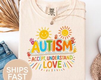 May include: Ivory-colored t-shirt with the word "AUTISM" in colorful letters, accompanied by the phrases "ACCEPT, UNDERSTAND, LOVE." The design includes smiling suns and handprints. The shirt is folded, with a necklace visible.
