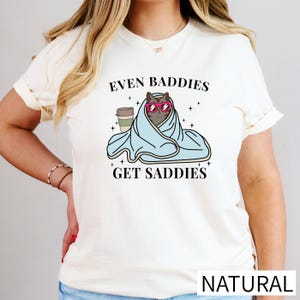 A woman wearing a white t-shirt with a graphic design of a cat wrapped in a blanket and wearing sunglasses, along with the text "Even Baddies Get Saddies".