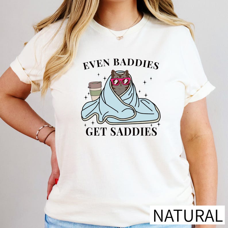 A woman wearing a white t-shirt with a graphic design of a cat wrapped in a blanket and wearing sunglasses, along with the text "Even Baddies Get Saddies".