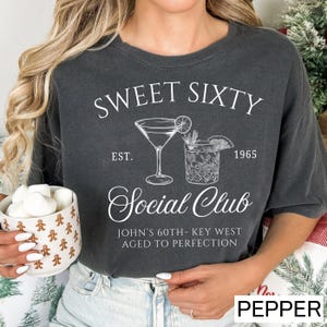 A woman wearing a black t-shirt with a graphic design that says "Sweet Sixty Social Club" and "John's 60th-Key West Aged to Perfection". She is holding a mug with marshmallows in it.