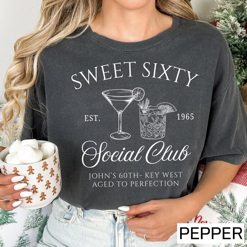 A woman wearing a black t-shirt with a graphic design that says "Sweet Sixty Social Club" and "John's 60th-Key West Aged to Perfection". She is holding a mug with marshmallows in it.