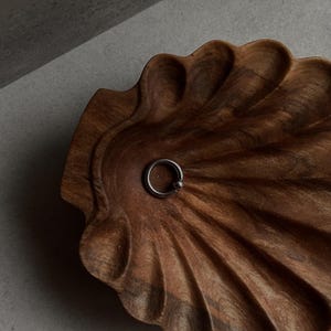 May include: A dark brown, wooden, shell-shaped bowl with a silver ring in the center. The bowl has a carved, textured surface and is set against a gray background. The ring is small and circular.