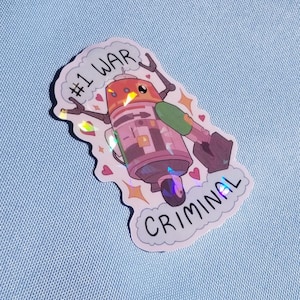 May include: Holographic sticker depicting a cartoon robot with the words "#1 WAR CRIMINAL". The robot is mainly pink, orange, and grey, with green and black details. The sticker has a white border and is decorated with hearts and stars.