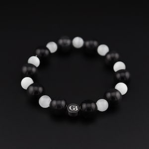 Puede incluir: Pulsera de cuentas negras y blancas con un dije plateado que dice "GB".