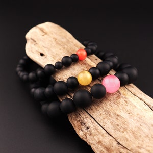 Puede incluir: Tres pulseras de cuentas negras con cuentas de colores. Las pulseras están apiladas sobre un trozo de madera a la deriva.