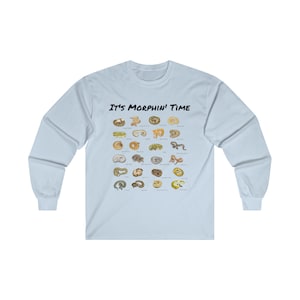 Puede incluir: Camiseta de manga larga azul claro con el texto "It's Morphin' Time" impreso en negro. La camiseta presenta un gráfico de varios morfos de serpiente con sus nombres enumerados debajo.
