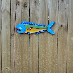 May include: A hand-painted wooden wall art piece shaped like a mahi-mahi fish. The fish features a vibrant blue dorsal fin, a yellow underbelly, and a green body with blue spots. It has a red mouth and a signature near the tail fin.
