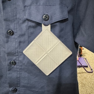 May include: A gray plastic badge holder with a diamond shape and a grid pattern. The badge holder is attached to a blue shirt with buttons.