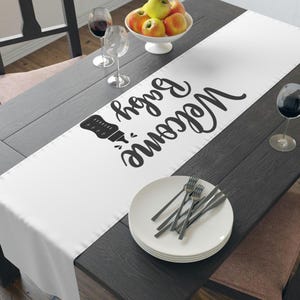 May include: A white table runner featuring the text "Welcome Baby" in black script and a baby bottle illustration. It's set on a dark wooden table, with plates, cutlery, and wine glasses also present.