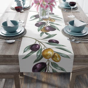 May include: A white table runner with a watercolor olive branch design. The runner features green leaves and purple and gold olives. The table is set with teal-colored bowls, plates, and wine glasses, creating an elegant dining scene.
