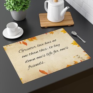 May include: A rectangular placemat with a vintage paper texture features the quote "Greater love has no one than this: to lay down one's life for one's friends." The placemat is decorated with autumn leaves.