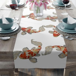 Puede incluir: Camino de mesa blanco con un patrón repetido de carpas koi en tonos rojos, naranjas y dorados. Las carpas koi tienen escamas detalladas y aletas fluidas. El camino de mesa está colocado sobre una mesa de madera, con cubiertos y un jarrón con flores rosas.