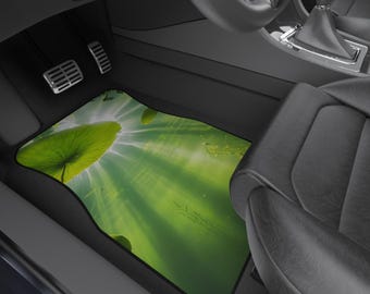 May include: Car floor mat featuring a vibrant green lotus leaf design with sun rays. The mat is rectangular with a black border, placed in a car interior with black seats and pedals.