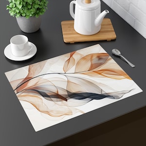 May include: A rectangular placemat with an abstract design of flowing, leaf-like shapes in shades of brown, orange, and gray against a white background. The placemat is on a dark surface, with a white teacup and saucer, a teapot, and a spoon nearby.