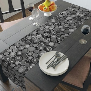 May include: A dark wooden dining table is set with a patterned runner featuring a black and white circular design. White plates with silverware, wine glasses, and a bowl of fruit complete the setting. The table is surrounded by chairs.