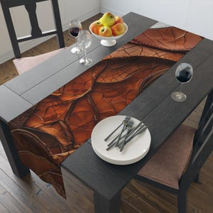 May include: A dining table set with a brown leather-textured table runner. The table is set with white plates, silverware, wine glasses, and a bowl of apples. The table and chairs are dark gray.