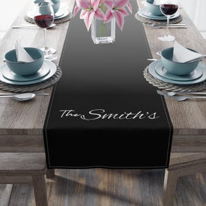 May include: A black table runner featuring a thin white border and the white cursive text The Smith's centered near the bottom edge. The runner is displayed on a wooden table set with blue bowls, white napkins, silverware, and wine glasses.