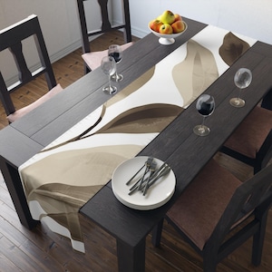 May include: A dining table set with a dark wood finish, featuring a decorative leaf-patterned table runner in shades of brown and beige. The table is set with white plates, silverware, wine glasses, and a bowl of fruit.