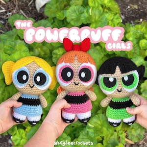 Puede incluir: Tres muñecas de ganchillo hechas a mano de Las Supernenas. Cada muñeca tiene un color de pelo único: amarillo, naranja y negro. Llevan vestidos en azul, rosa y verde, con rayas negras. Las muñecas tienen ojos grandes y caras sonrientes.