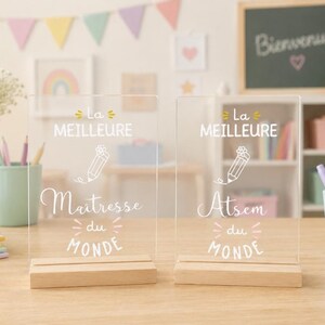 Peut inclure: Deux plaques rectangulaires en acrylique transparent avec des socles en bois. Chaque plaque présente un texte blanc et un dessin de crayon. La plaque de gauche indique La MEILLEURE Maitresse du MONDE et celle de droite La MEILLEURE Atsem du MONDE.