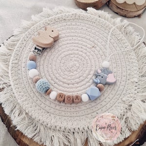 Peut inclure: Un hochet pour bébé bleu clair et blanc avec des perles en bois et un charme en bois d'éléphant et d'écureuil. Le hochet est sur une surface en crochet blanc avec le nom "NOA" écrit en lettres en bois.