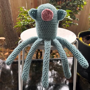 K&ouml;nnte beinhalten: Handgefertigtes H&auml;keltier in Form eines Oktopus in T&uuml;rkis und Rosa. Der Oktopus hat einen runden Kopf mit zwei kleinen, ohrenartigen Anh&auml;ngseln und einem rosa Kreis in der Mitte. Acht lange Tentakel erstrecken sich vom K&ouml;rper. Das Spielzeug steht auf einem wei&szlig;en Metallhocker.