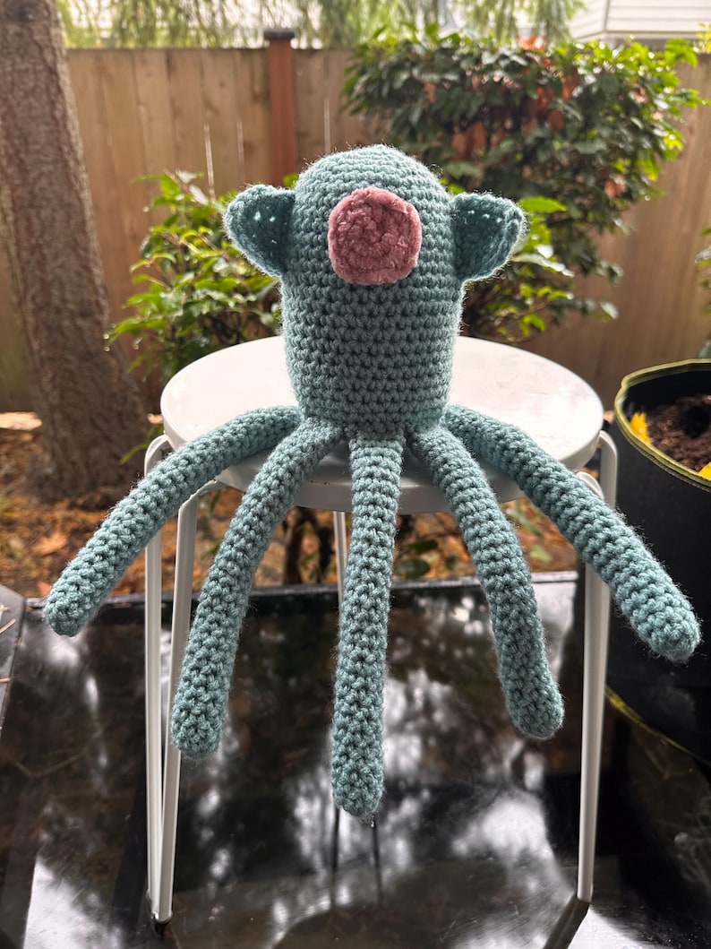 K&ouml;nnte beinhalten: Handgefertigtes H&auml;keltier in Form eines Oktopus in T&uuml;rkis und Rosa. Der Oktopus hat einen runden Kopf mit zwei kleinen, ohrenartigen Anh&auml;ngseln und einem rosa Kreis in der Mitte. Acht lange Tentakel erstrecken sich vom K&ouml;rper. Das Spielzeug steht auf einem wei&szlig;en Metallhocker.