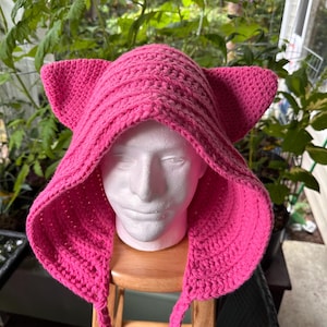 May include: A bright pink crocheted hood featuring two prominent, triangular cat-like ears on top. The hood is constructed with a textured yarn stitch pattern and includes two long, braided yarn ties hanging down from the front edges of the garment.
