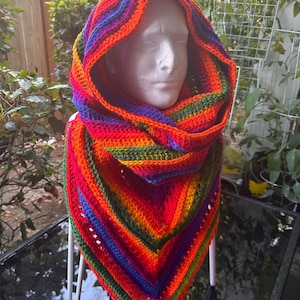 May include: A vibrant, handmade crochet hood and scarf set. The hood and scarf are a spectrum of colors, including red, orange, yellow, green, blue, and purple. The hood is draped over a white mannequin head, showcasing the design and texture of the crochet.