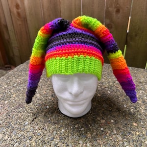 May include: A colorful crocheted hat with long, pointed ear flaps. The hat is striped with black, purple, pink, orange, yellow, and green yarn.