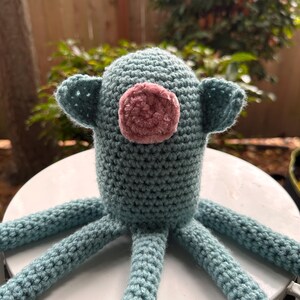 K&ouml;nnte beinhalten: Handgefertigtes H&auml;keltier in Form eines Oktopus in T&uuml;rkis und Rosa. Der Oktopus hat einen runden Kopf mit kleinen Ohren und einen rosa, flauschigen Kreis im Gesicht. Sechs t&uuml;rkisfarbene Tentakel erstrecken sich vom K&ouml;rper. Das Spielzeug liegt auf einer wei&szlig;en Oberfl&auml;che.