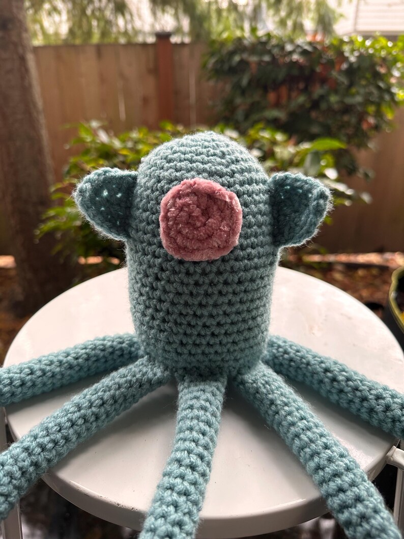 K&ouml;nnte beinhalten: Handgefertigtes H&auml;keltier in Form eines Oktopus in T&uuml;rkis und Rosa. Der Oktopus hat einen runden Kopf mit kleinen Ohren und einen rosa, flauschigen Kreis im Gesicht. Sechs t&uuml;rkisfarbene Tentakel erstrecken sich vom K&ouml;rper. Das Spielzeug liegt auf einer wei&szlig;en Oberfl&auml;che.