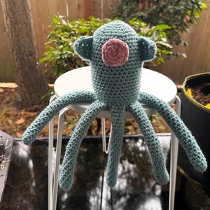 K&ouml;nnte beinhalten: Handgefertigte H&auml;kel-Oktopus-Puppe in T&uuml;rkis und Rosa. Die Puppe hat einen runden Kopf mit kleinen ohrartigen Vorspr&uuml;ngen und einem rosa kreisf&ouml;rmigen Gesicht. Sechs lange Tentakel erstrecken sich vom K&ouml;rper. Die Puppe sitzt auf einem wei&szlig;en Hocker.