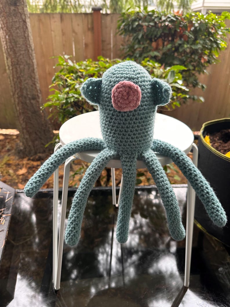 K&ouml;nnte beinhalten: Handgefertigte H&auml;kel-Oktopus-Puppe in T&uuml;rkis und Rosa. Die Puppe hat einen runden Kopf mit kleinen ohrartigen Vorspr&uuml;ngen und einem rosa kreisf&ouml;rmigen Gesicht. Sechs lange Tentakel erstrecken sich vom K&ouml;rper. Die Puppe sitzt auf einem wei&szlig;en Hocker.