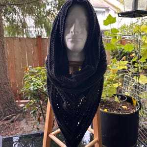 May include: A black crocheted hooded scarf displayed on a mannequin head. The scarf is triangular, with a hood that frames the face. The crochet pattern is visible, and the scarf is draped over a wooden stool. The background includes greenery and a wooden fence.