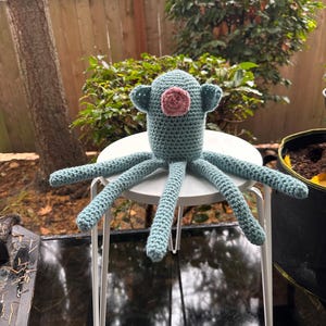 K&ouml;nnte beinhalten: Ein handgefertigter H&auml;kel-Oktopus in T&uuml;rkis- und Rosat&ouml;nen sitzt auf einem kleinen wei&szlig;en Tisch. Der Oktopus hat acht lange, strukturierte Tentakel und einen runden Kopf mit kleinen, ohrenartigen Anh&auml;ngseln. Der Hintergrund zeigt Gr&uuml;n und einen Holzzaun.