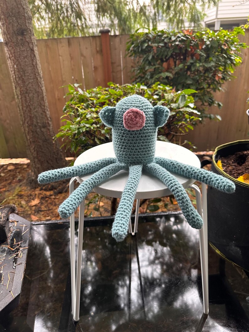 K&ouml;nnte beinhalten: Ein handgefertigter H&auml;kel-Oktopus in T&uuml;rkis- und Rosat&ouml;nen sitzt auf einem kleinen wei&szlig;en Tisch. Der Oktopus hat acht lange, strukturierte Tentakel und einen runden Kopf mit kleinen, ohrenartigen Anh&auml;ngseln. Der Hintergrund zeigt Gr&uuml;n und einen Holzzaun.